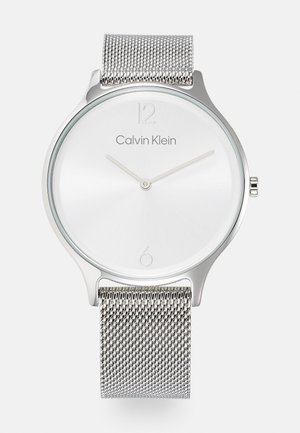 Sølv mesh rem ur med et minimalistisk rundt urværk. Hvid skive med sølv time markeringer og "Calvin Klein" logo ved kl. 12.