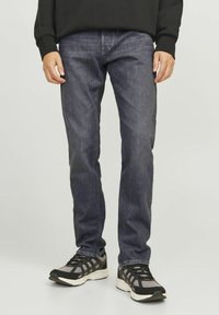 Mand iført mørkegrå slim-fit jeans og sorte og grå sportssko, stående mod en ensartet lys baggrund.