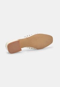 Zapato de tacón alto color crema con suela de goma texturizada, que presenta detalles recortados en la parte superior y un diseño de tacón cuadrado.