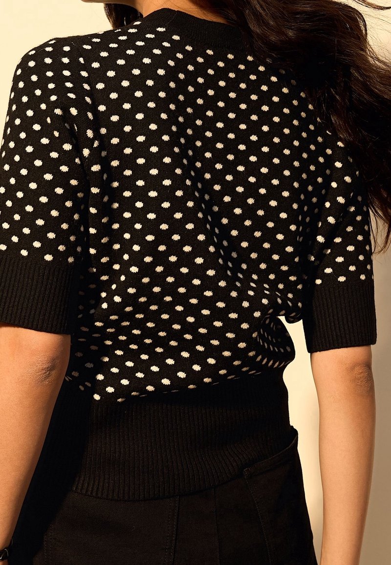Femme portant un pull noir à manches courtes avec des pois blancs, poignets et taille côtelés, vue de dos avec des cheveux foncés lâchés.