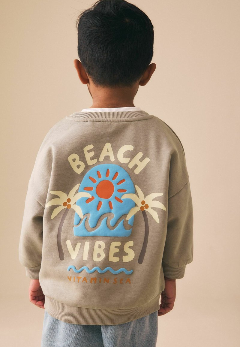 Dieťa otočené chrbtom, oblečené v béžovej mikine s textom „Beach Vibes“, obrazcom slnka, palmových stromov a vĺn na chrbte.