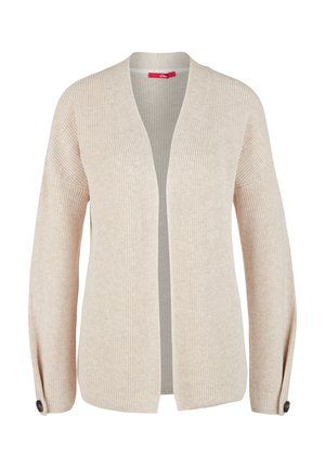 Femme portant un gilet beige, un chemisier à fleurs vert foncé, un pantalon vert olive, une ceinture noire et des baskets blanches, debout avec une main dans la poche.