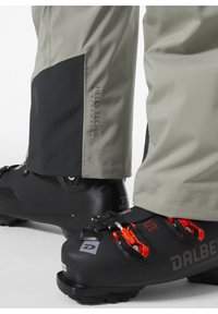 Zwarte skibots met oranje accenten en metalen sluitingen, gecombineerd met grijze waterdichte broeken met een zwart paneel en HELLY TECH branding.