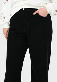 Pantalons en denim noir avec une coupe droite, poches avant et fermeture à bouton en métal. La texture est lisse avec un effet de décoloration minimal.