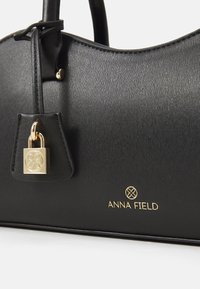 Bolso negro hecho de material sintético texturizado. Cuenta con herrajes dorados, un detalle de candado y el logo "ANNA FIELD" en dorado en la parte inferior.