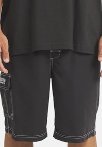 Mustat lyhyet housut kevyestä materiaalista, joissa on valkoiset ompeluyksityiskohdat. Sivutasku logopatchilla. Leikkaus hiukan polven yläpuolelle.