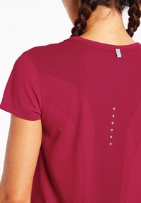 Kvinna med flätat hår som bär en kortärmad, ventilerande sporttröja i maroon med reflexpunkter på övre ryggen.