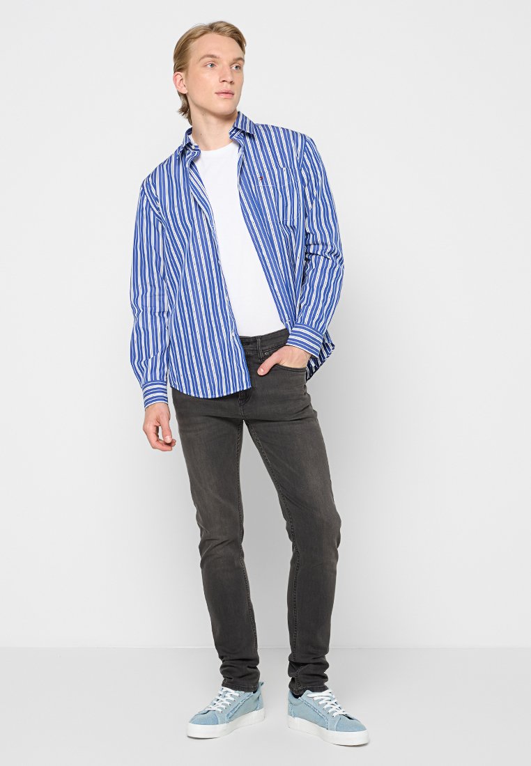 Giovane in piedi con una mano in tasca, indossa una camicia a righe blu e bianche sopra una maglietta bianca, jeans neri e sneakers azzurre chiare.