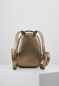 Metallisch bronzefarbener Rucksack mit runder Form, glatter Oberfläche und zwei verstellbaren Trägern. Verfügt über eine kleine Seitentasche zur Aufbewahrung.