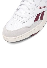 Biele tenisky s hladkým koženým zvrškom a semišovými akcentmi v svetlo sivej farbe, červenými pruhmi, perforovanou špičkou a textúrovanou podrážkou.