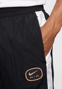 Pantalones cortos de nailon negros con paneles laterales blancos, que presentan un parche redondo bordado con "Nike AIR" en naranja. Cintura elástica, detalle de bolsillo con cremallera.