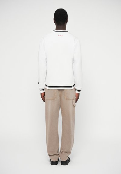 Λευκό φούτερ με μαύρο περίγραμμα, με κόκκινο κείμενο στην πλάτη. Συνδυάζεται με ανοιχτό μπεζ cargo παντελόνι και μαύρα παπούτσια.