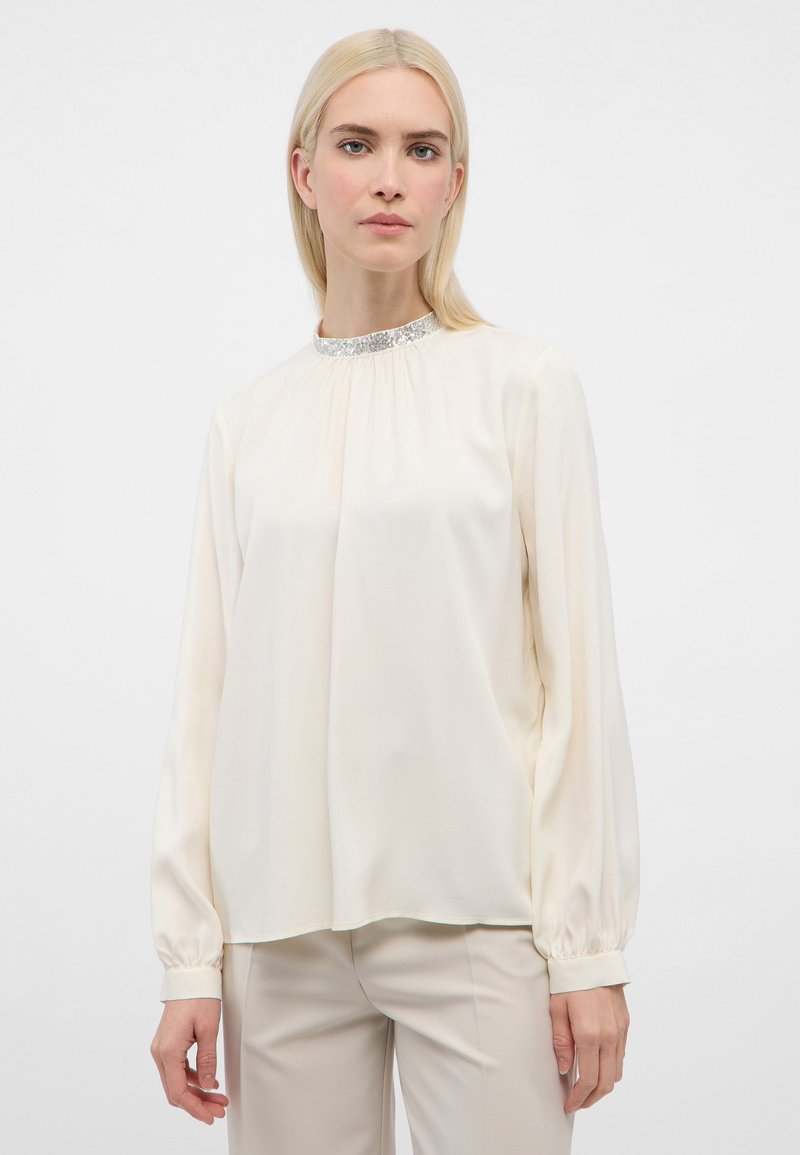 Langærmet bluse i cremet silke med en rynket hals og sølvglitrende palietkant. Har elastiske manchetter og en løs pasform. Kombineret med lyse bukser.