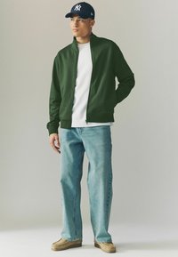 Veste zippée verte sur un t-shirt blanc, associée à un jean large bleu clair et des mocassins marron, couronnée d'une casquette navy.