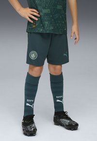 Grön idrottströja med geometriska mönster, matchande shorts, mörkgröna strumpor med texten "CITY" och svarta fotbollsskor med gröna detaljer.