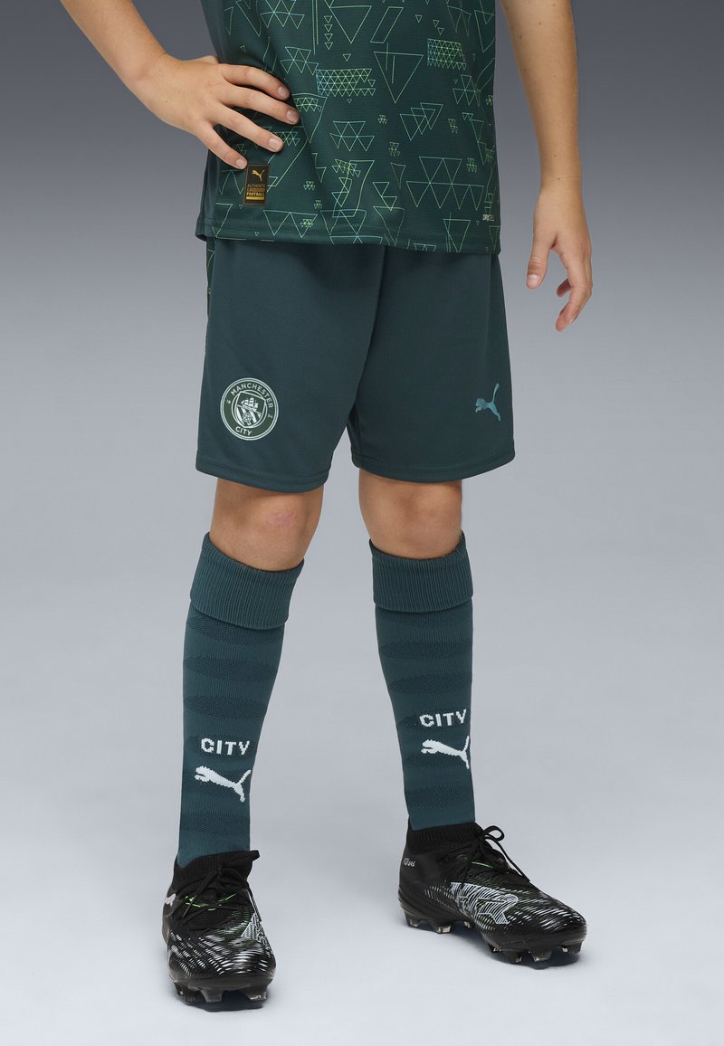 Grön idrottströja med geometriska mönster, matchande shorts, mörkgröna strumpor med texten "CITY" och svarta fotbollsskor med gröna detaljer.