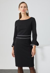 Schwarzes Kleid mit langen Ärmeln, ausgestellten Manschetten und weißem Besatz an der Taille. Glattes Material, knielanges Design, minimalistischer Stil.