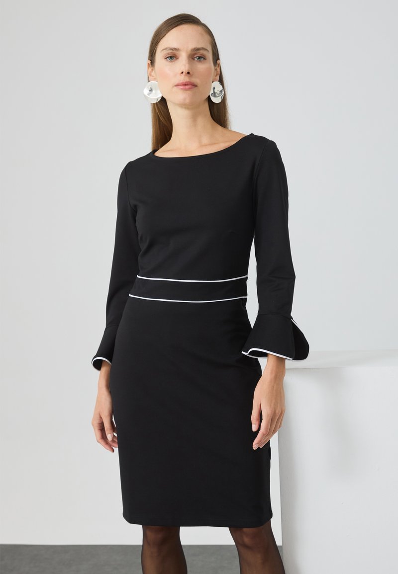 Schwarzes Kleid mit langen Ärmeln, ausgestellten Manschetten und weißem Besatz an der Taille. Glattes Material, knielanges Design, minimalistischer Stil.