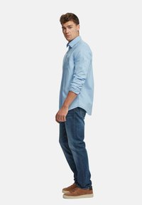 Helles blaues Hemd mit aufgekrempelten Ärmeln, kombiniert mit dunklen Jeans und braunen Sneakers. Das Modell steht seitlich vor einem weißen Hintergrund.