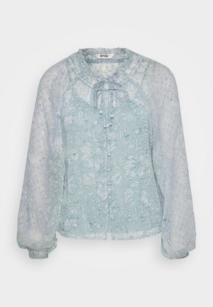 Blouse bleu clair en tissu transparent avec des motifs floraux, encolure froncée, fermeture à nouer et manches longues bouffantes.