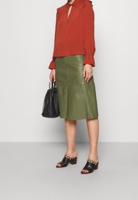 Rostfarbige Bluse mit Rüschen, grüner Leder-A-Linien-Rock und schwarze Absatzmules mit goldenen Akzenten. Hält eine schwarze Bucket Bag.