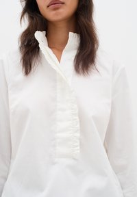 Vit bomullskjorta med rynkad placket, knäppning och mandarin-krage. Slät textur med en ren, minimalistisk design.