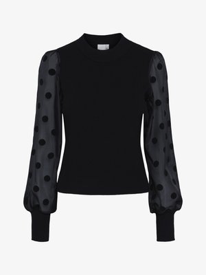 Must sõleb väike must sviiter, millel on läbipaistvad puff-siidised pikad varrukad, millel on suured mustad täpid ning ribad käeotsadel ja kaeluses.