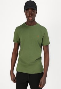 T-shirt verde-oliva com decote redondo e mangas dobradas, apresentando um pequeno logo bordado laranja "II" no peito. Boné preto usado.