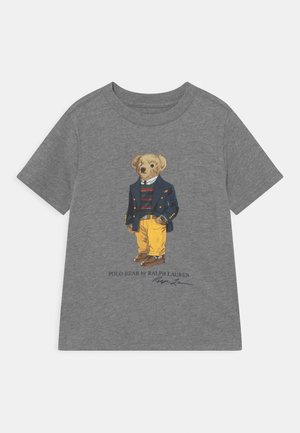 Grå T-shirt med illustration af en bjørn iført en marineblå blazer, stribet sweater og gule bukser med brune sko og signaturdetalje.
