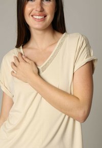 Haut beige à manches courtes avec un col en V, comprenant des détails en dentelle texturée le long de l'encolure. Tissu doux avec une coupe décontractée.