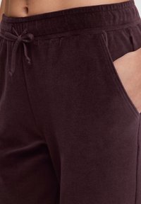 Close-up van een persoon in donkerpaarse sweatpants met een trekkoord in de taille en een hand in de voorzak.