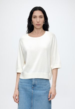 Mujer con cabello largo y oscuro, vestida con una camiseta blanca de manga tres cuartos y una falda vaquera azul, de pie frente a un fondo liso y claro.