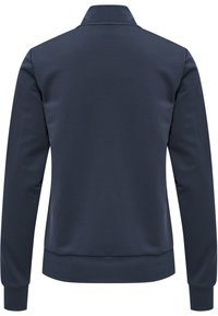 Marinblå zip-up jacka med ståkrage, långa ärmar och ribbade muddar och avslut; slätt texturerat material, minimalistisk design.