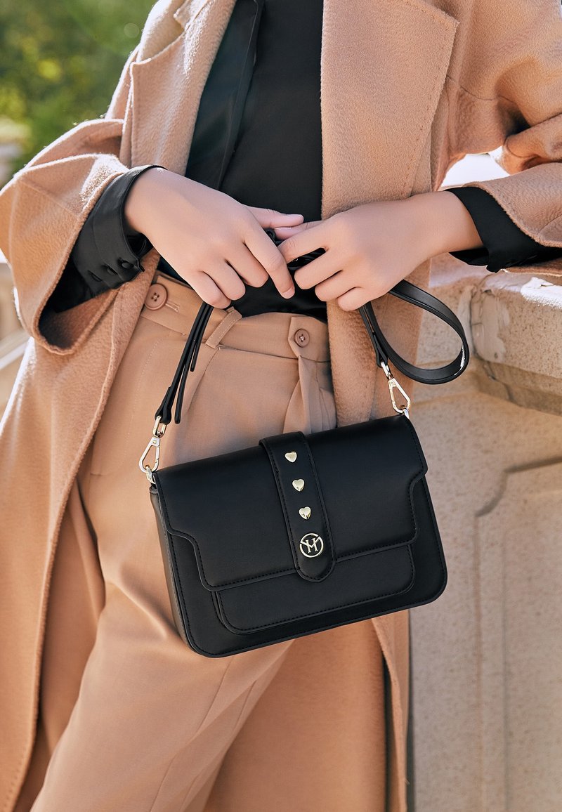 Personne portant un manteau et un pantalon beige tient un sac à main noir avec des clous dorés en forme de cœur, debout à côté d’une rambarde en pierre à l’extérieur.