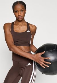 Καφέ αθλητικό σουτιέν με λευκές ρίγες και λογότυπο, σετ με κολάν ψηλόμεσο που ταιριάζει, κρατώντας μια μαύρη ιατρική μπάλα.