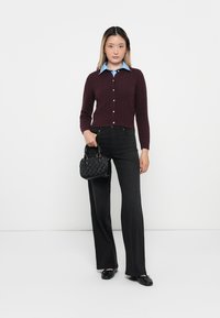 Cardigan bordeaux avec fermeture à bouton, associé à une chemise à col bleu clair, un pantalon noir évasé et un petit sac à main noir.