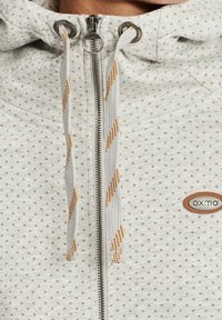 Sweat à capuche gris clair avec fermeture éclair, orné de petits points marron. Comprend une capuche à cordon avec des accents orange et blanc, ainsi qu'un patch logo sur la poitrine.