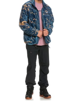 Giacca in pile blu con motivi astratti beige, chiusura con cerniera e colletto alto, abbinata a pantaloni cargo neri e stivali neri.