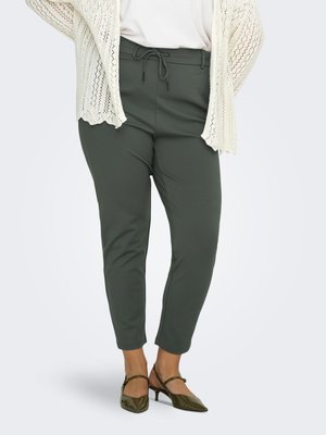 Žena u tamnozelenim uskim hlačama s vezicom u struku, bijeloj majici, kremasto pletenom kardiganu i maslinastozelenim cipelama s niskom petom.