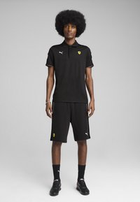 Camiseta polo negra con cuello y un sutil ribete, combinada con pantalones cortos negros. Presenta el logo de Ferrari y la marca Puma. Se lleva con zapatos y calcetines negros.