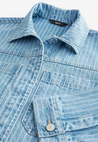 Camisa de mezclilla azul claro con rayas verticales, que cuenta con un cuello tipo spread, botón en la parte delantera y dos bolsillos en el pecho con acentos metálicos.