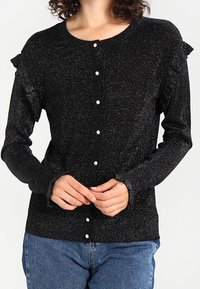 Svart knapp-up kofta med glittrande textur, volanger vid axlarna och pärlknappar. Bärs över en ljusblå denimunderdel.