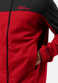 Veste rouge et noire avec fermeture éclair à l'avant, fabriquée en tissu doux. Présente un logo sur la poitrine, des poignets élastiques et un réglage de taille avec cordon de serrage.