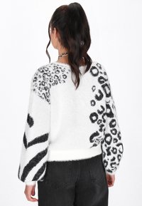 Weißer, fusseliger Pullover mit schwarzen Leopardenmuster auf den Ärmeln und dem Rücken, mit geripptem Saum und Bündchen. Kombiniert mit schwarzen Jeans.