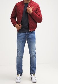 Chaqueta bomber acolchada roja con puños acanalados, combinada con un suéter de cuello redondo navy y jeans desgastados de color azul claro. Zapatillas blancas completan el look.