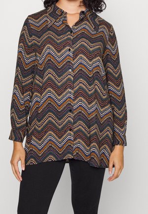 Chemise à manches longues avec boutons, présentant un motif chevron en zigzag orange, marine, blanc et beige, portée avec un pantalon noir.