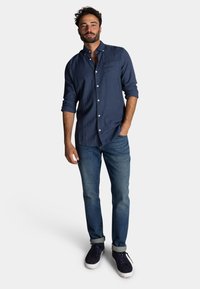 Marineblauw overhemd met knoopsluiting en borstzak, opgerolde mouwen, gecombineerd met blauwe jeans en donkergekleurde sneakers, tegen een witte achtergrond.