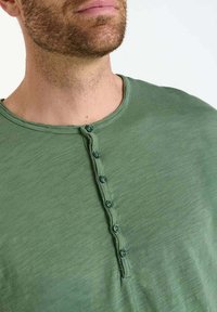 Green, short-sleeve henley shirt made of soft fabric, featuring a crew neck and six button placket with matching buttons, subtly textured.