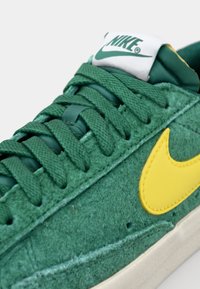 Zeleni semšasti copat z teksturirano zgornjo plastjo, rumenim Nike Swooshom, zelenimi vezalkami in belim oznakovnim jezikom, ki vsebuje logotip Nike.