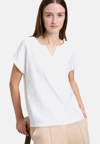 Blusa bianca con maniche corte e scollo a V, caratterizzata da un motivo floreale testurizzato. Il tessuto morbido cade in modo ampio, abbinata a pantaloni beige chiaro.